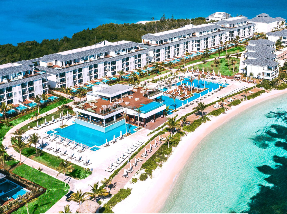 Aerial view of a luxury beachfront resort with multiple pools, sandy beach, and ocean shoreline in the background.