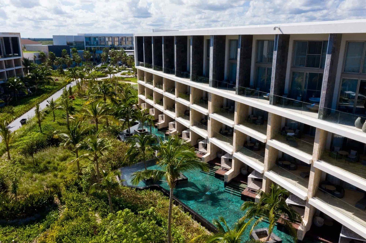 View of a modern hotel with multiple floors and balconies, surrounded by lush greenery, palm trees, and a swimming pool area.