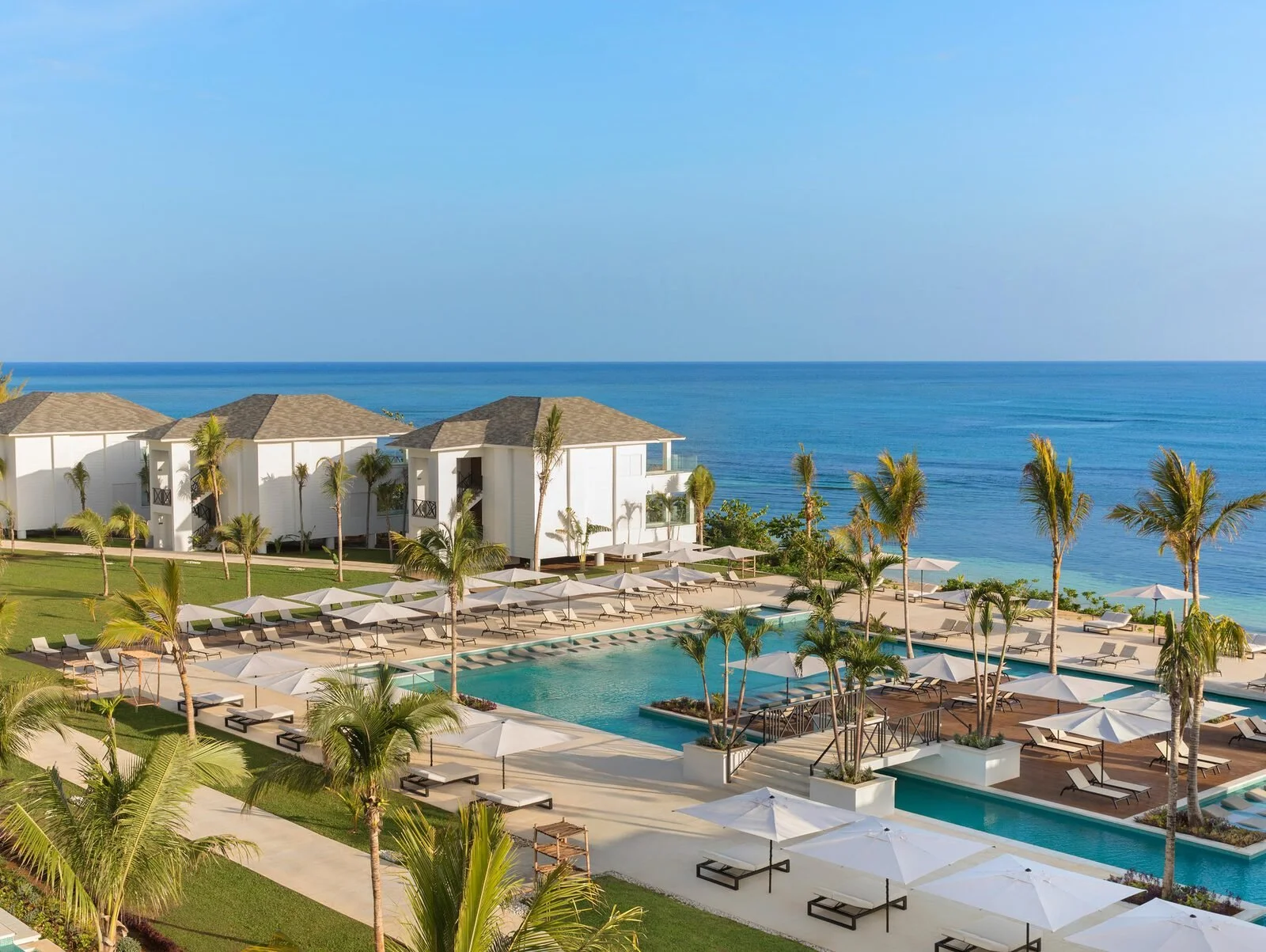 Luxury seaside resort with white buildings, swimming pools, beach chairs, umbrellas, and palm trees overlooking the ocean.