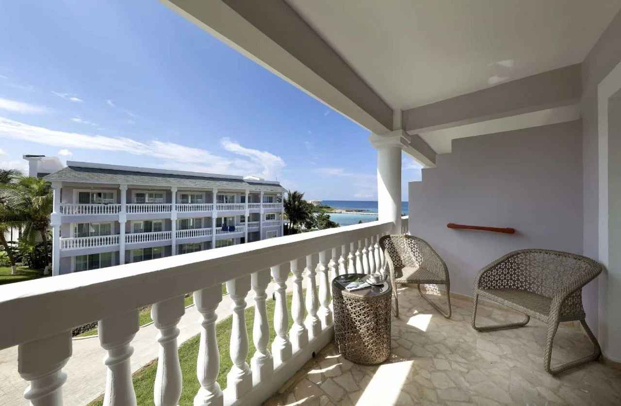 A balcony with outdoor chairs and a small table overlooking a white building with balconies and palm trees, with the ocean and blue sky in the background.