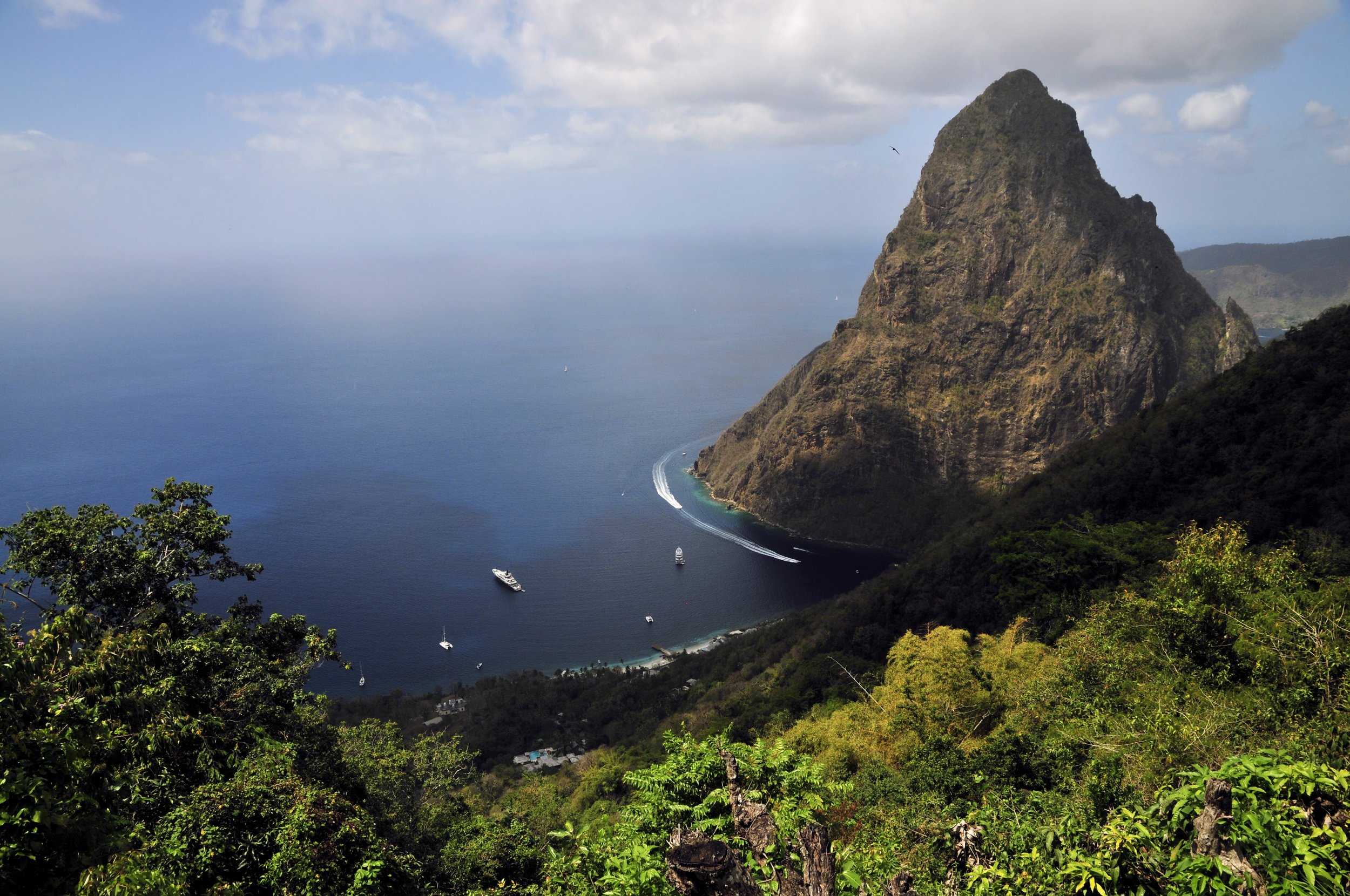 A view of a large mountain rising from the ocean with boats near its base and lush green trees in the foreground.
