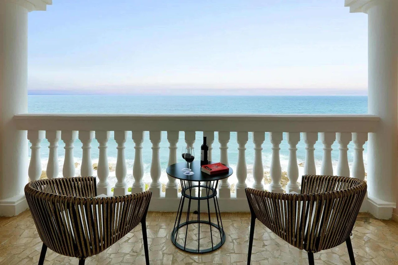 A balcony with two wicker chairs and a small black side table holding a glass of red wine, a wine bottle, and a red book, overlooking the ocean.