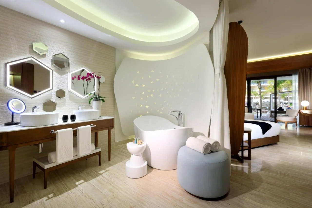Modern hotel bathroom with a white freestanding bathtub, a vanity with two sinks, decorative mirrors, a potted orchid, and soft lighting. A curtain separates the bathroom from a bedroom with a large bed, outdoor view, and seating area.