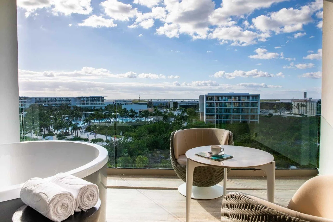 Hotel balcony with a view of a blue sky, clouds, and modern building structures in the distance. There is a round table with a coffee cup, a towel on a chair, and a bathtub on the balcony.