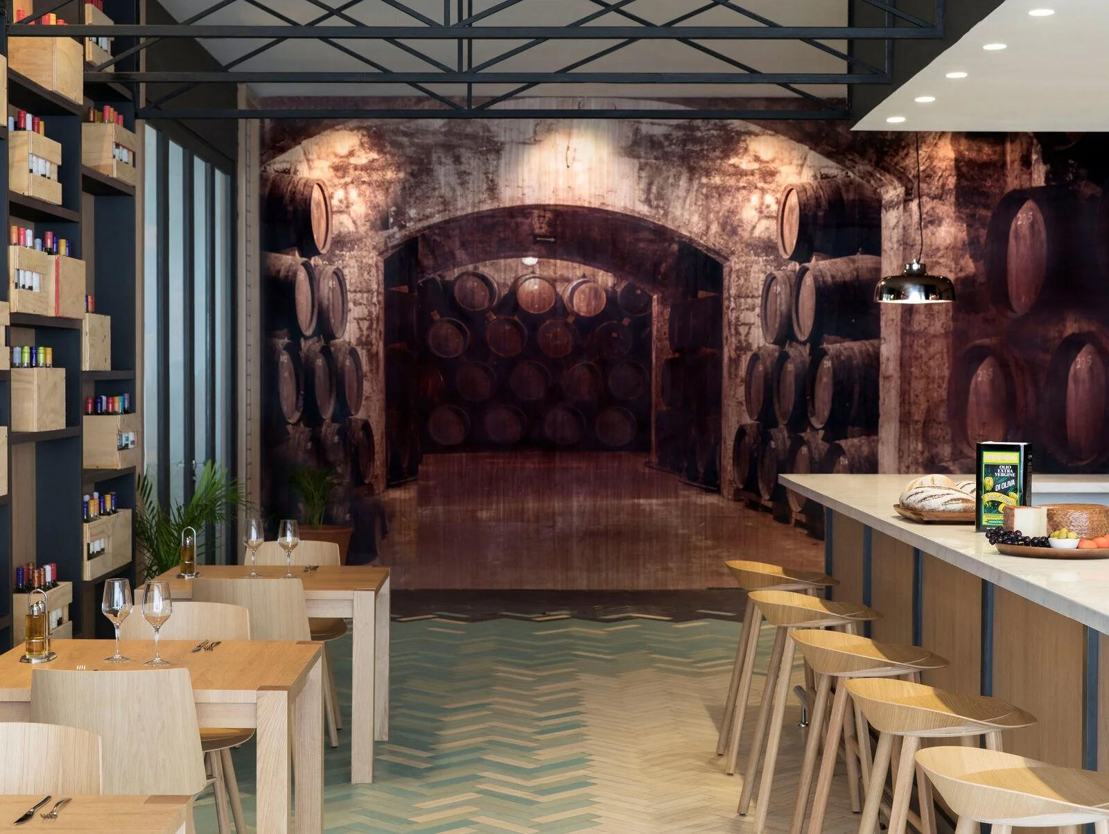 Interior of a modern restaurant with light wood tables and chairs, wine glasses, a bar counter with cheese and grapes, and a large wall mural of wine barrels in a cellar.