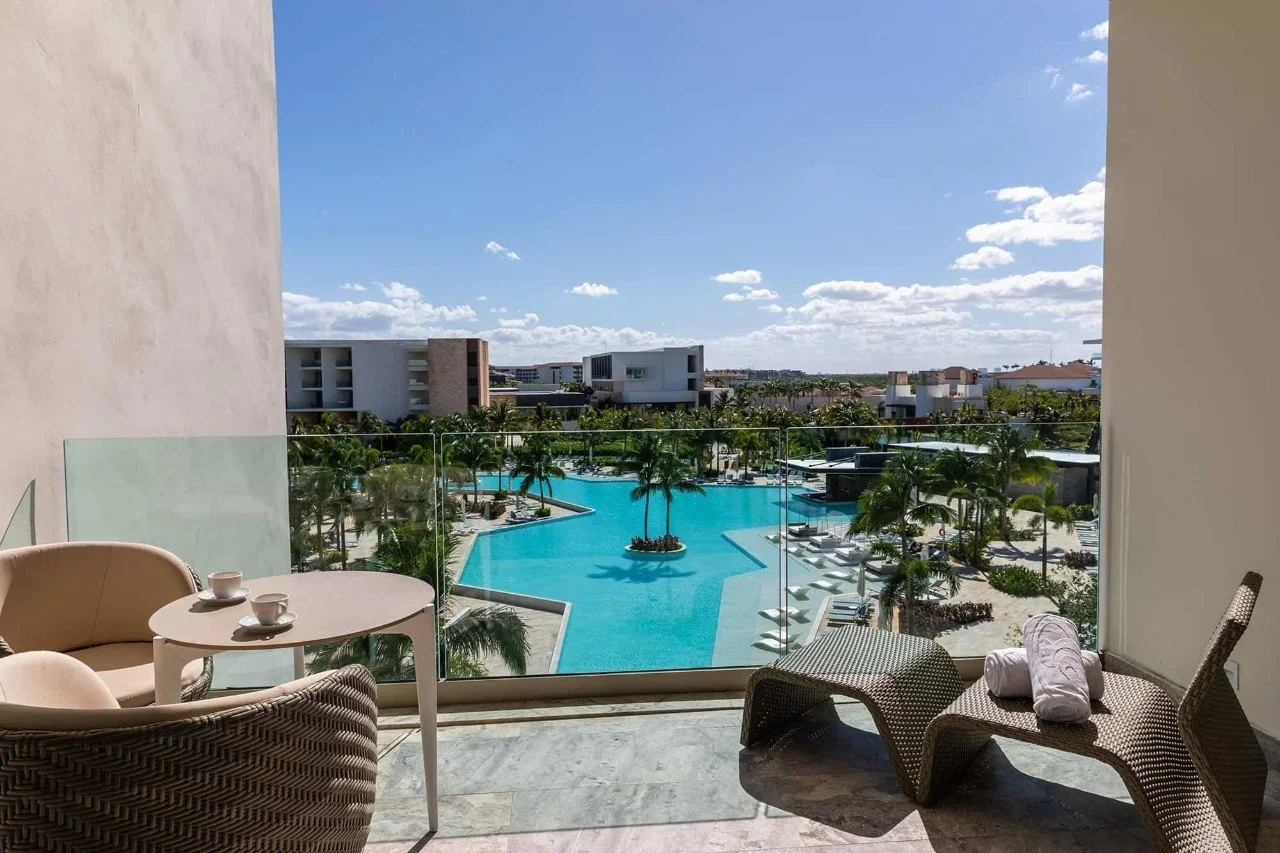 Balcony view of a resort pool area with lounge chairs, palm trees, and modern white buildings under a blue sky with scattered clouds.