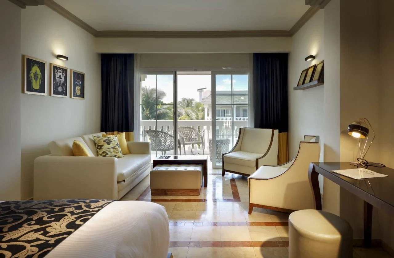 Interior of a hotel room with a bed in the foreground, a sitting area with a white sofa, armchair, and small table, large sliding glass door leading to a balcony with chairs, and decor including framed pictures on the wall and a desk with a lamp.