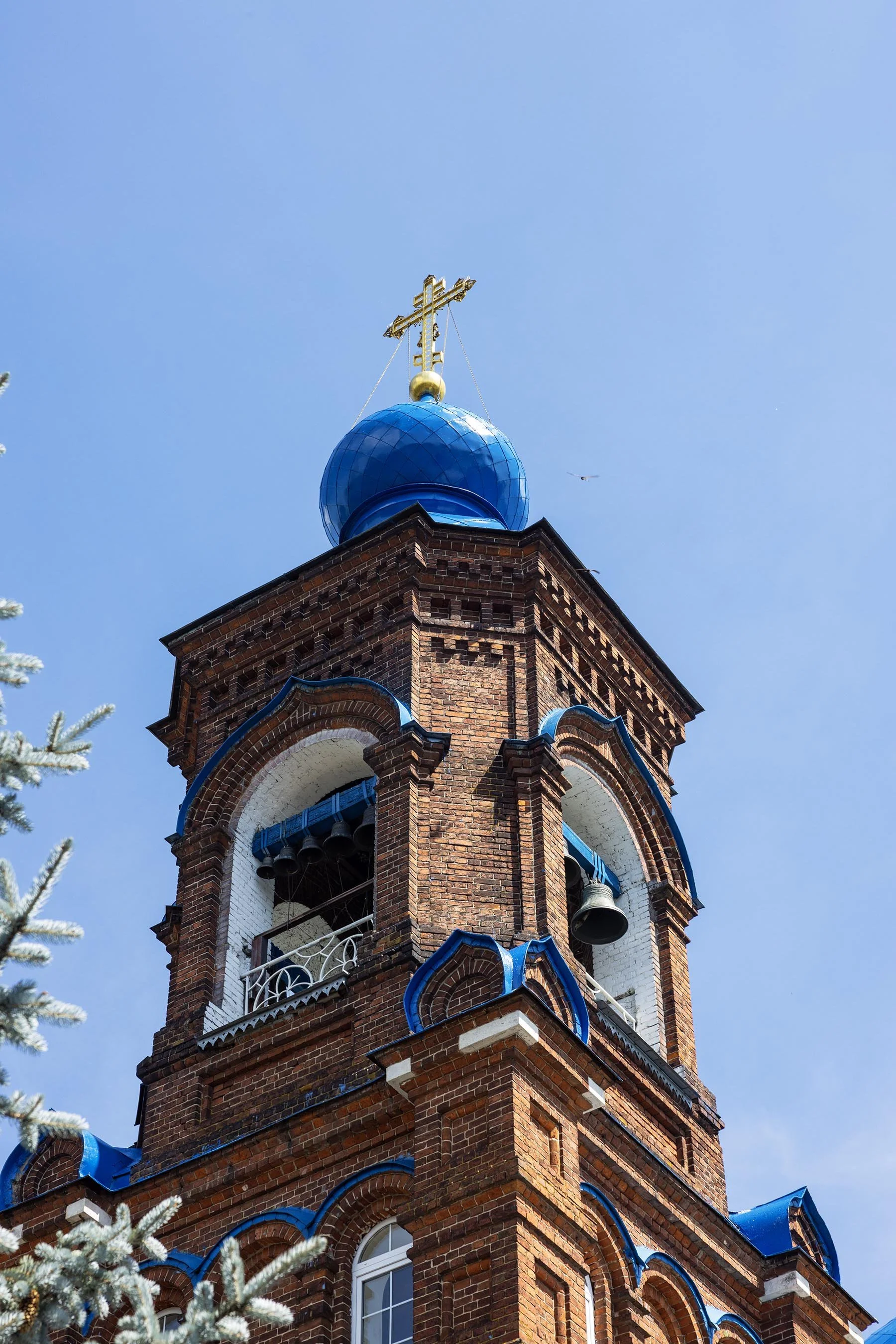 A church steeple with a gold cross on top, a blue onion-shaped dome, and brick architecture against a clear blue sky.