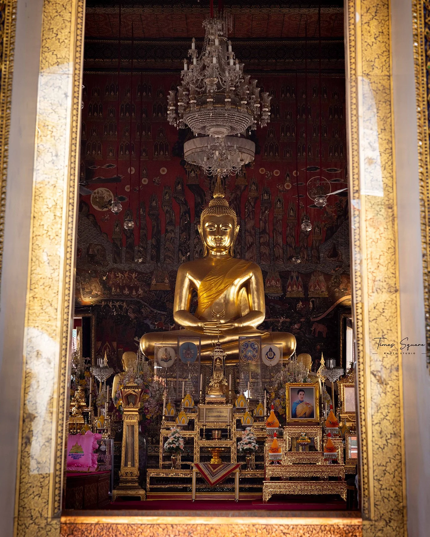 A large gold Buddha statue seated in meditation inside a Buddhist temple, framed by a decorative doorway. The temple interior features intricate artwork, a chandelier, and various religious artifacts and portraits.