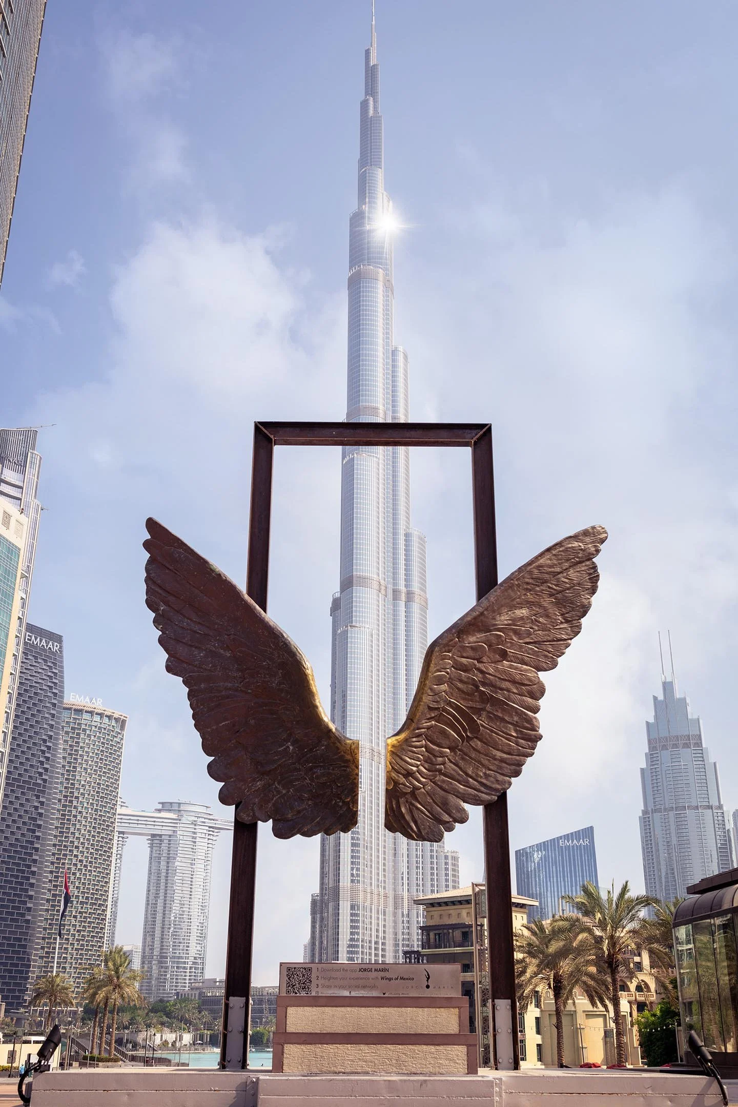 A bronze sculpture with large wings and an open frame in front of the Burj Khalifa skyscraper in Dubai, with surrounding high-rise buildings and palm trees.