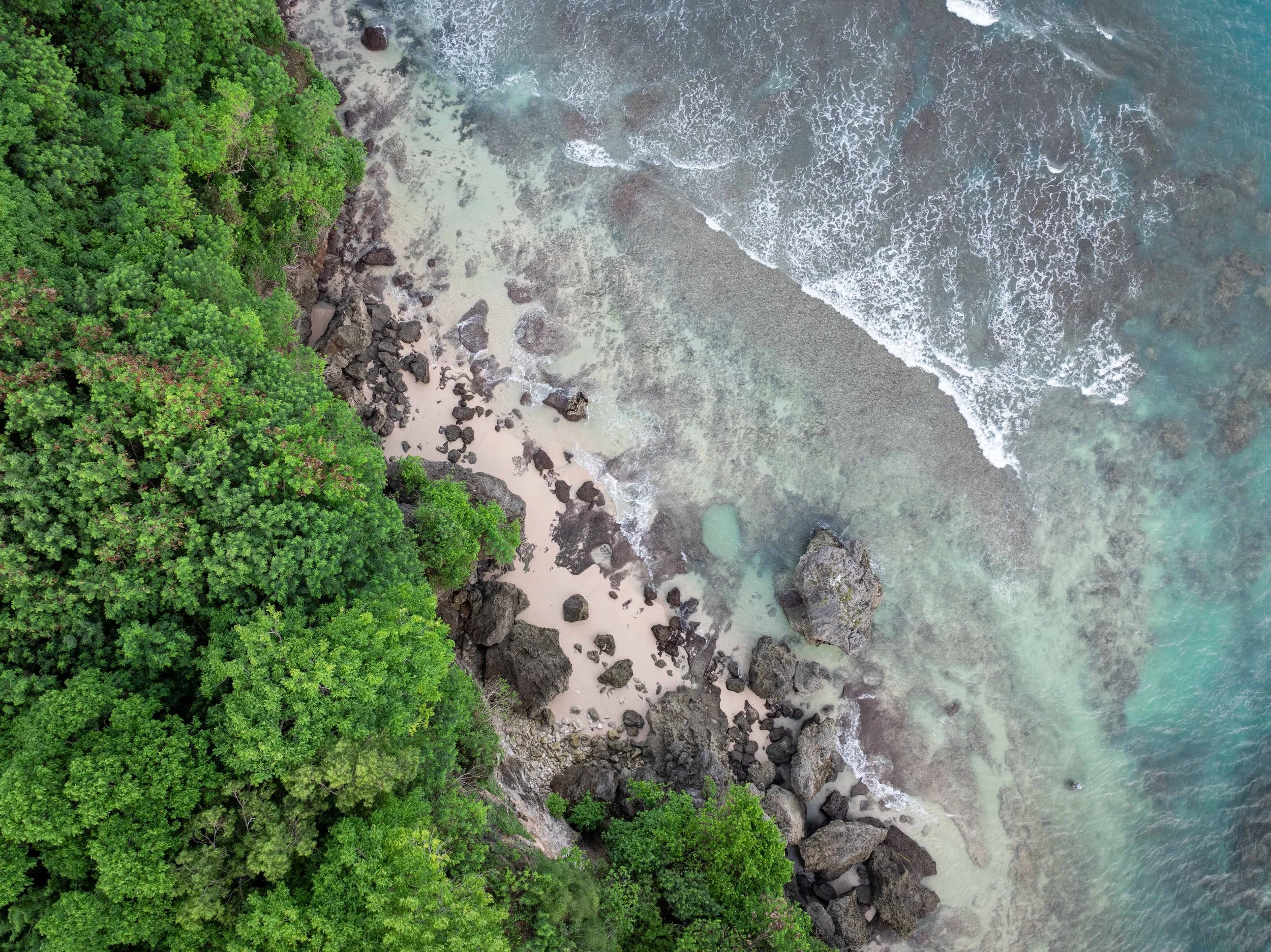 An aerial view of a beach with rocks, sand, ocean water, and dense green trees along the shoreline.