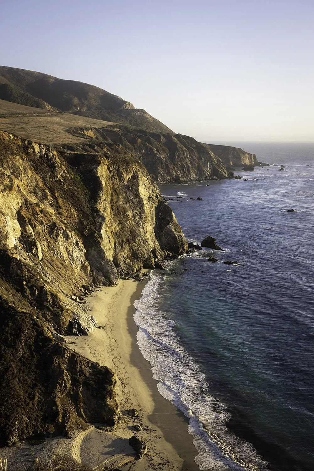 Scenic view of cliffs along a beach with ocean waves in the foreground and a clear sky.