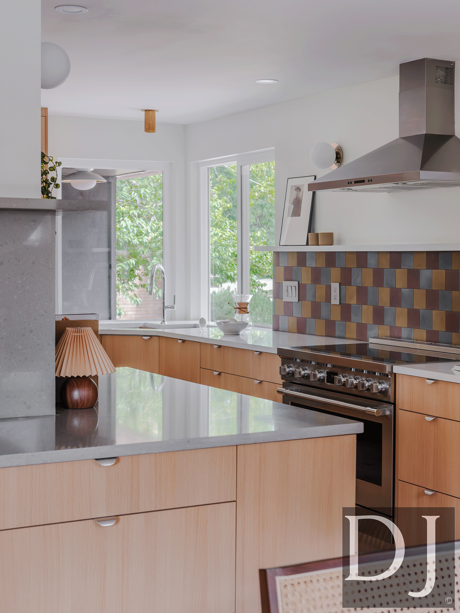 Modern kitchen with light wood cabinets, stainless steel oven and range, mosaic tile backsplash, large window with green trees outside, and various decorative items on the counter.