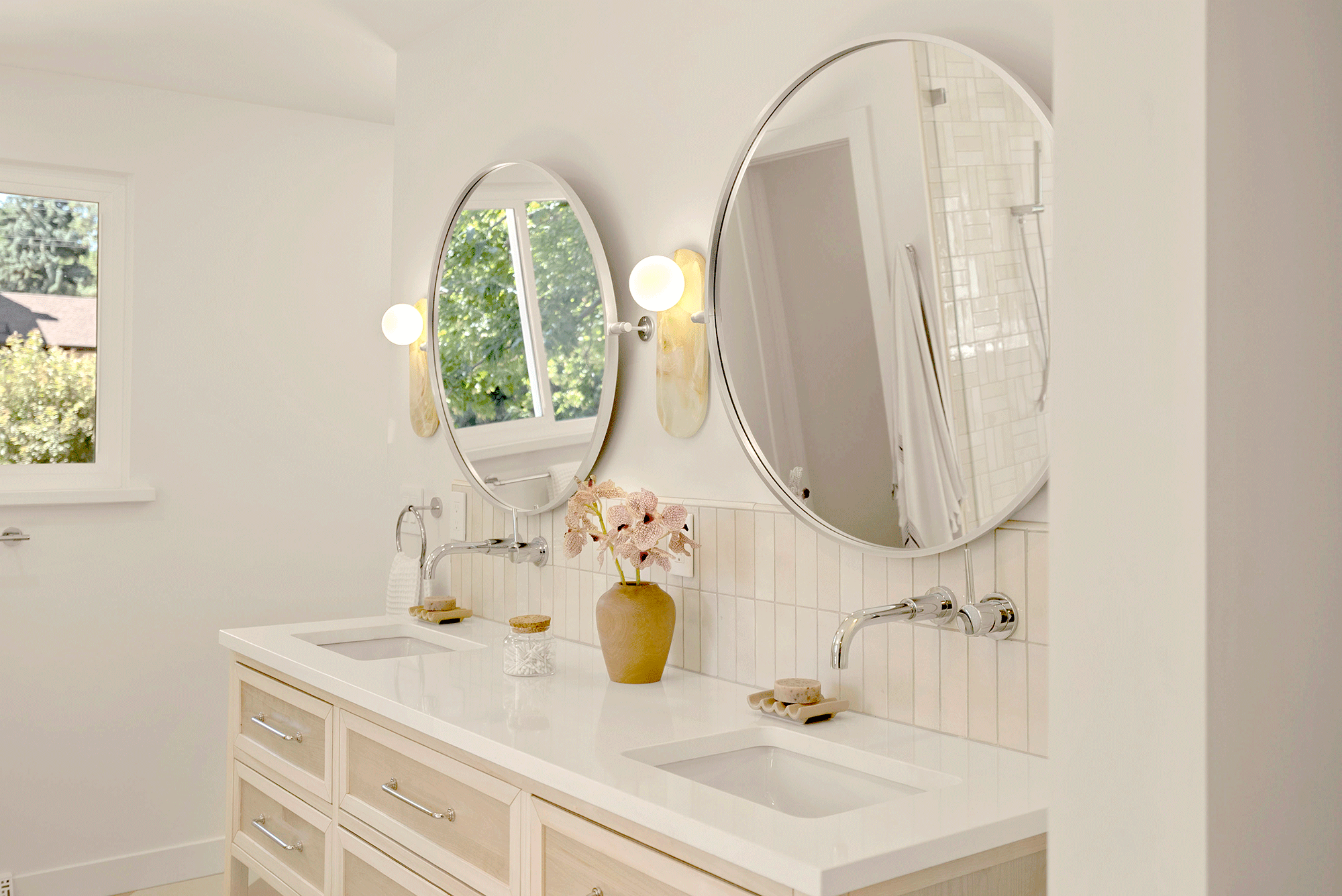 Bathroom vanity with dual sinks, large round mirrors, natural light from window, flowers in a vase, decorative jars, and shower area with glass enclosure in background.