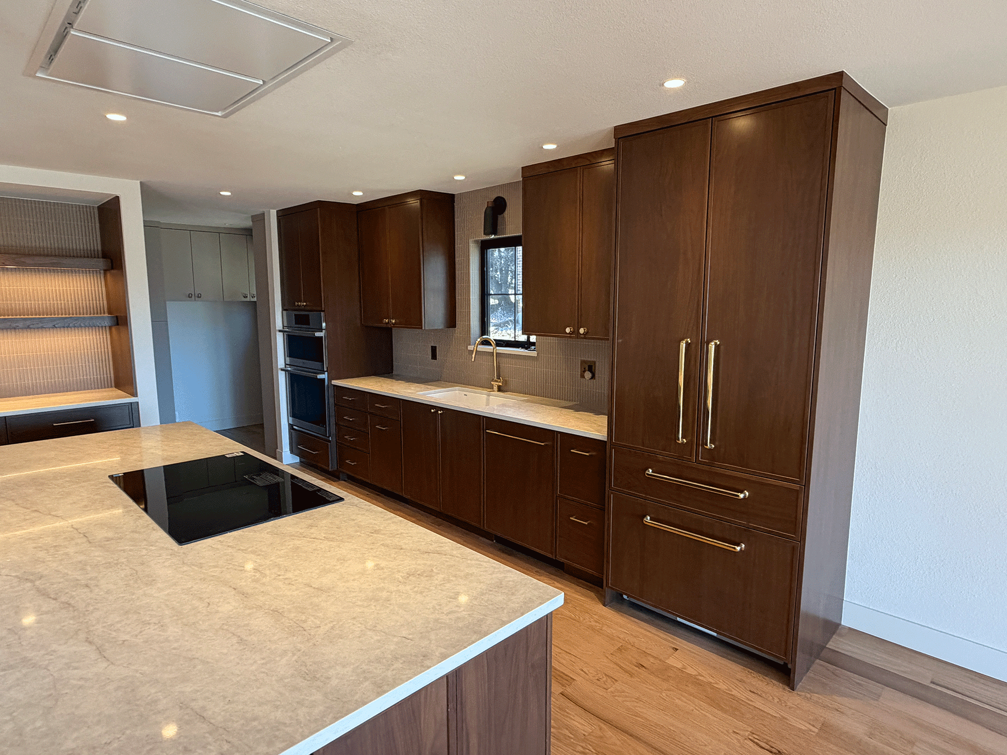 A modern kitchen with brown wooden cabinets, a cream-colored marble countertop, and a black electric stovetop. There is a window with a view outside and recessed ceiling lights illuminating the space.