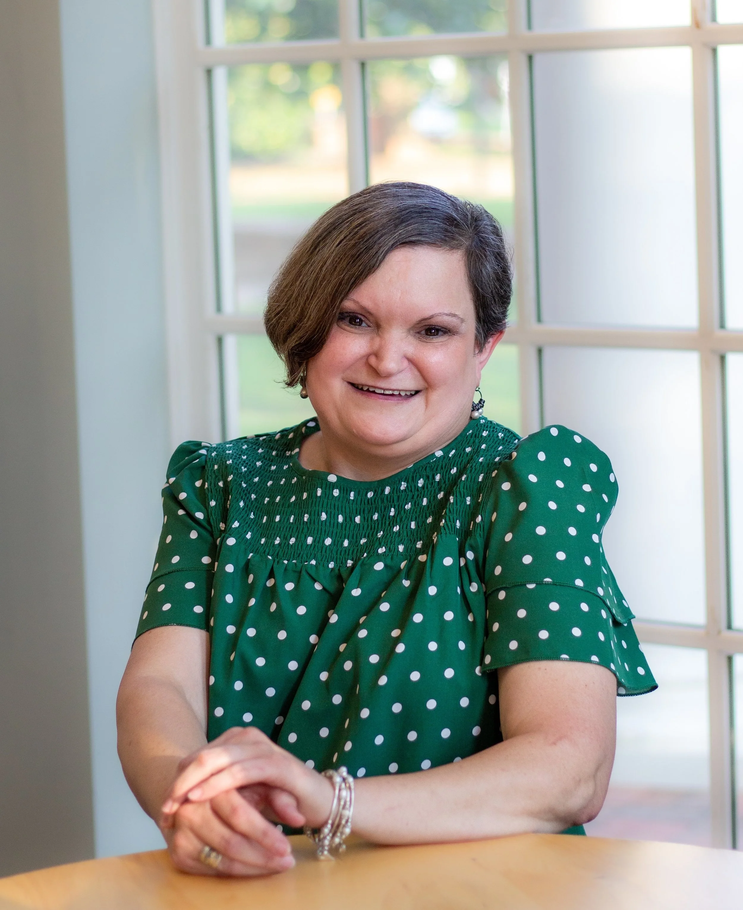 A woman with short brown hair wearing a green polka dot dress, smiling, sitting at a wooden table in front of a large window with greenery outside.