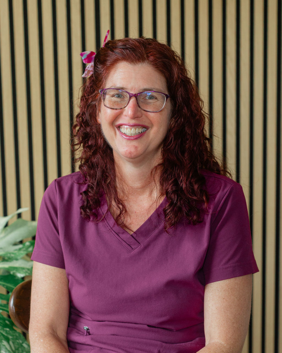 A woman with curly red hair wearing glasses and a black top, smiling at the camera in an indoor setting.
