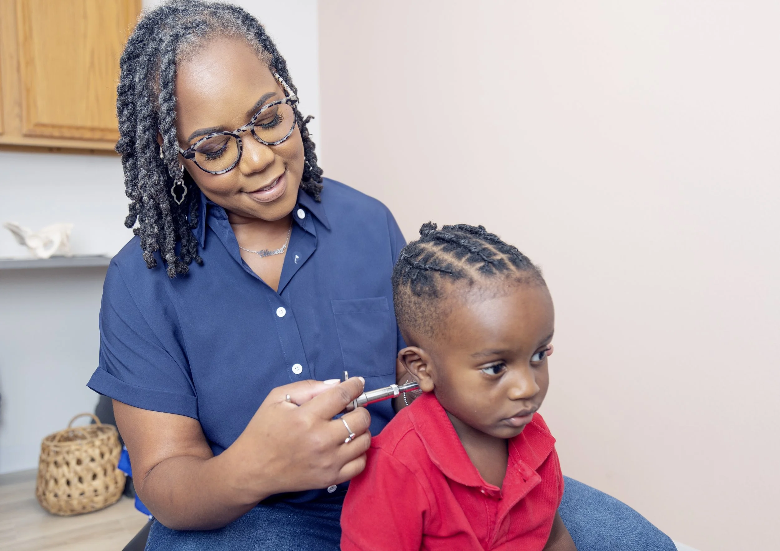 A woman with glasses and curly hair giving a young boy a medical ear examination with an otoscope.