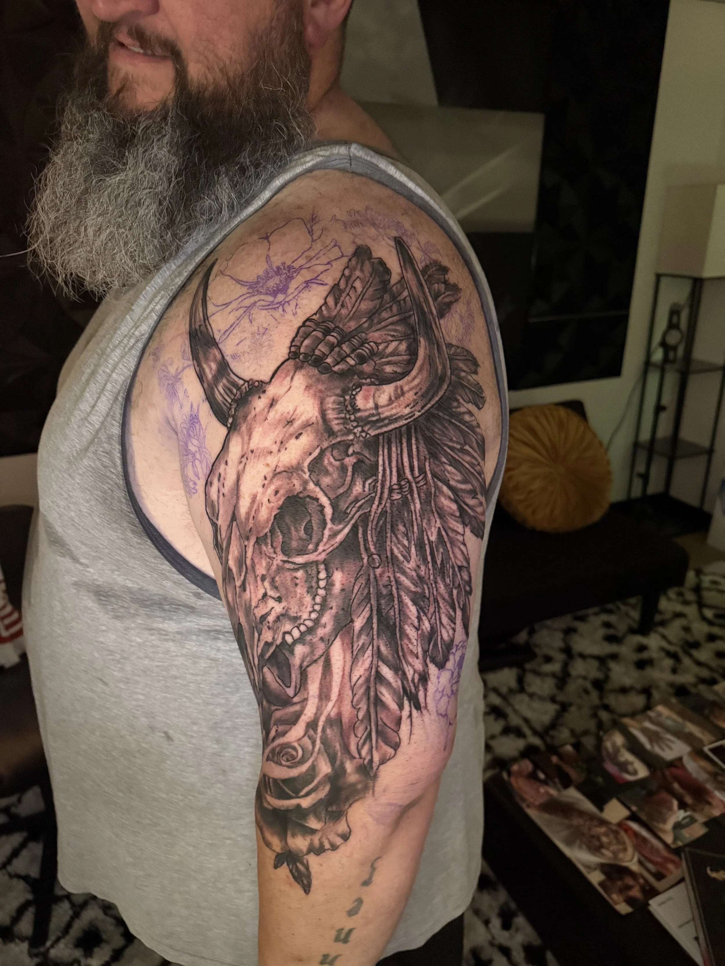 A man with a gray beard displaying a detailed black and gray tattoo of a deer skull with feathers on his upper arm.