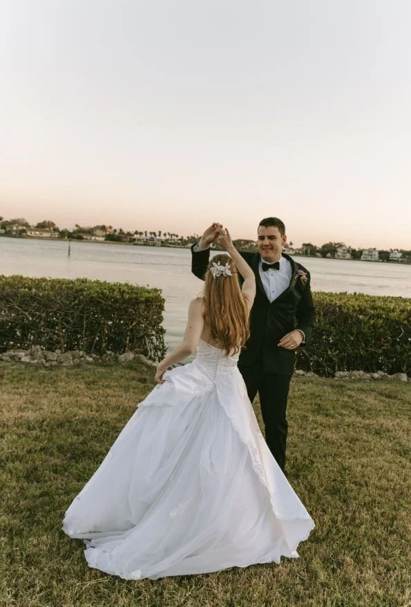 Groom twirling his bride by her hand at a tropical waterfront at golden hour sunset, with her white ballgown spinning and a flower hair clip visible — a romantic and playful couples portrait from a Tampa Bay wedding