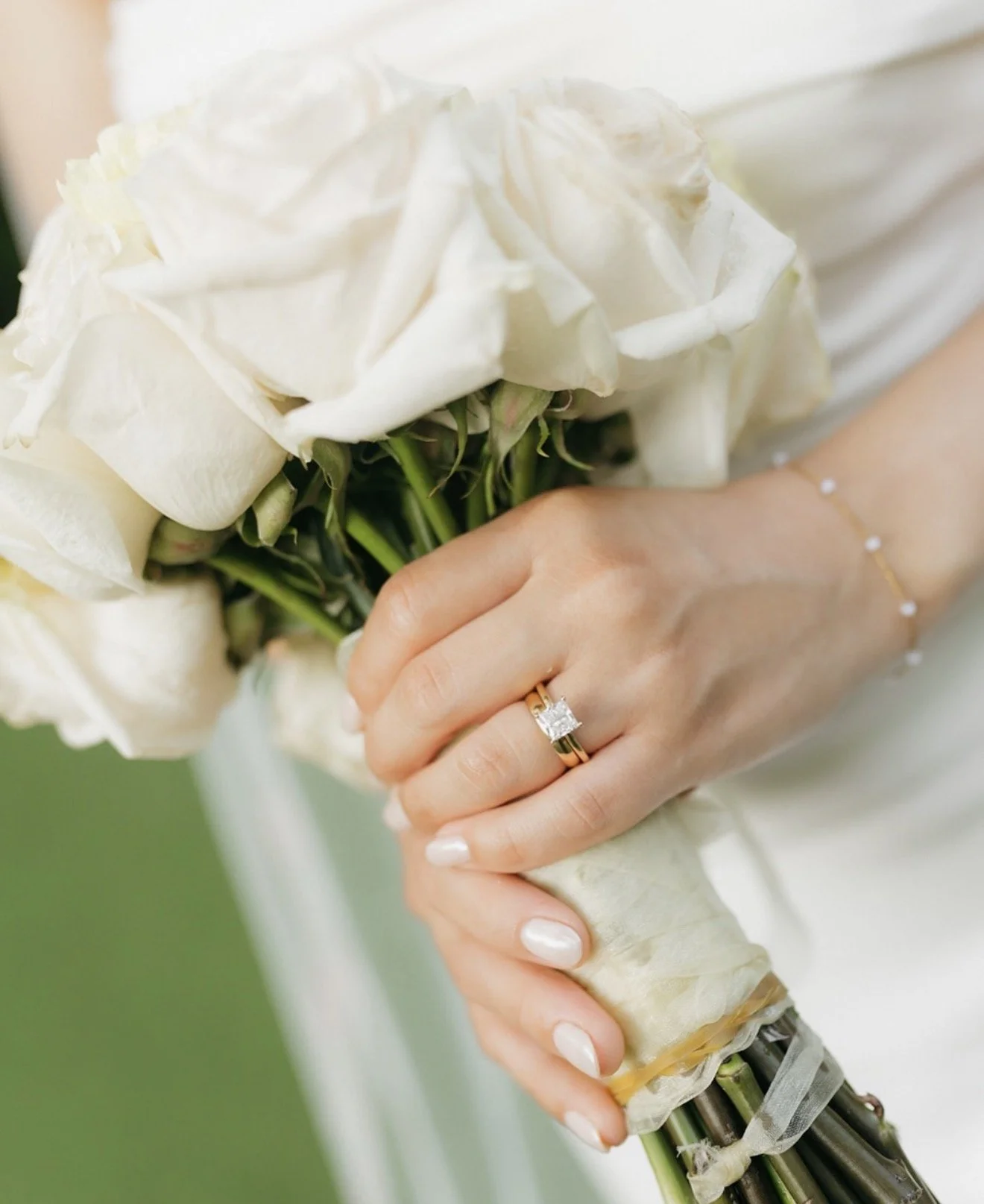 Close-up of bride's hand holding a white rose bouquet with a gold princess-cut diamond engagement ring and gold wedding band, soft blush nails and pearl bracelet, at Tampa Sunken Gardens wedding by Peony Wedding Co.