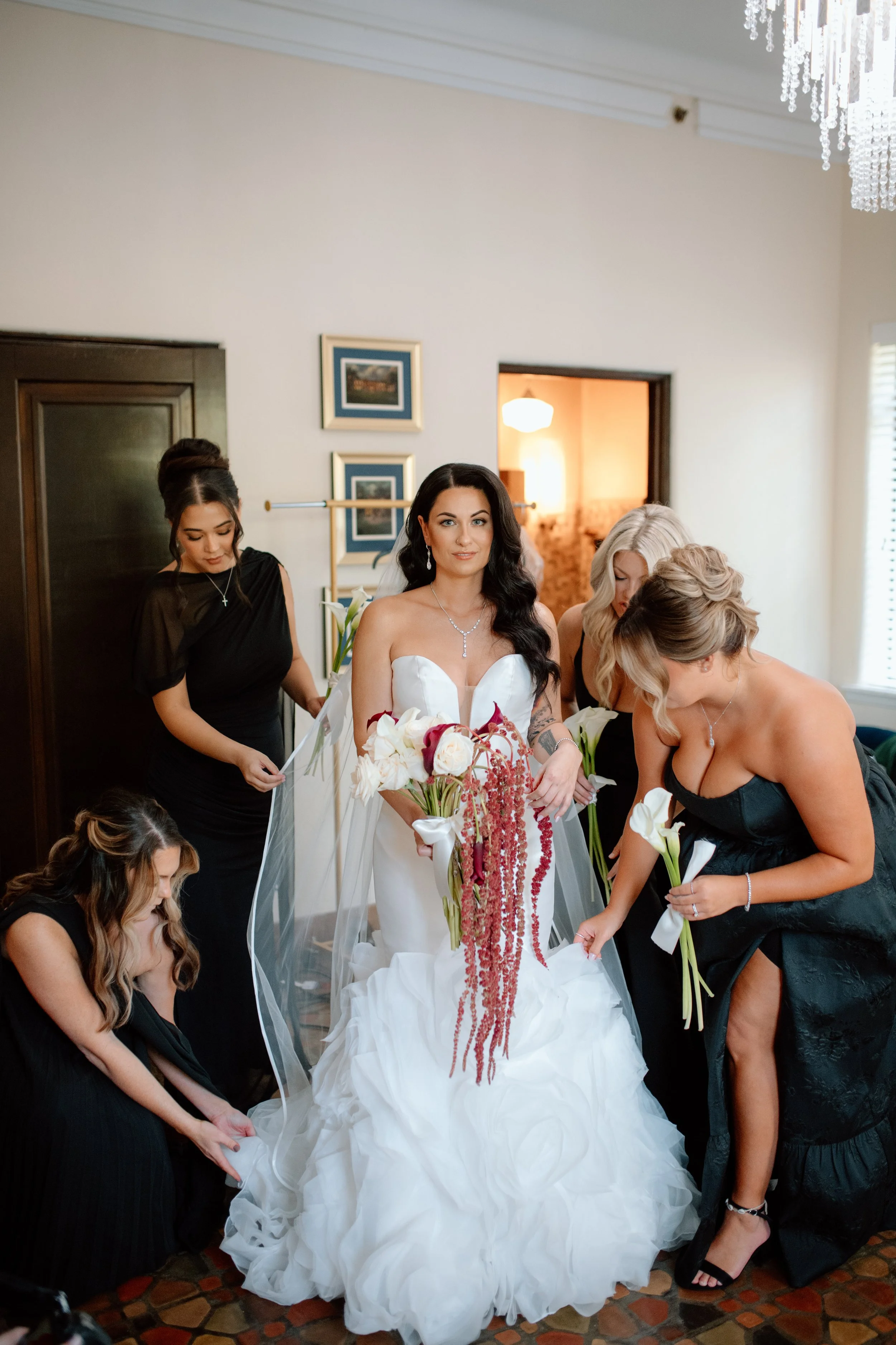 Bride in a white strapless gown holding a cascading red and white bouquet, surrounded by bridesmaids in black dresses helping her get ready indoors.