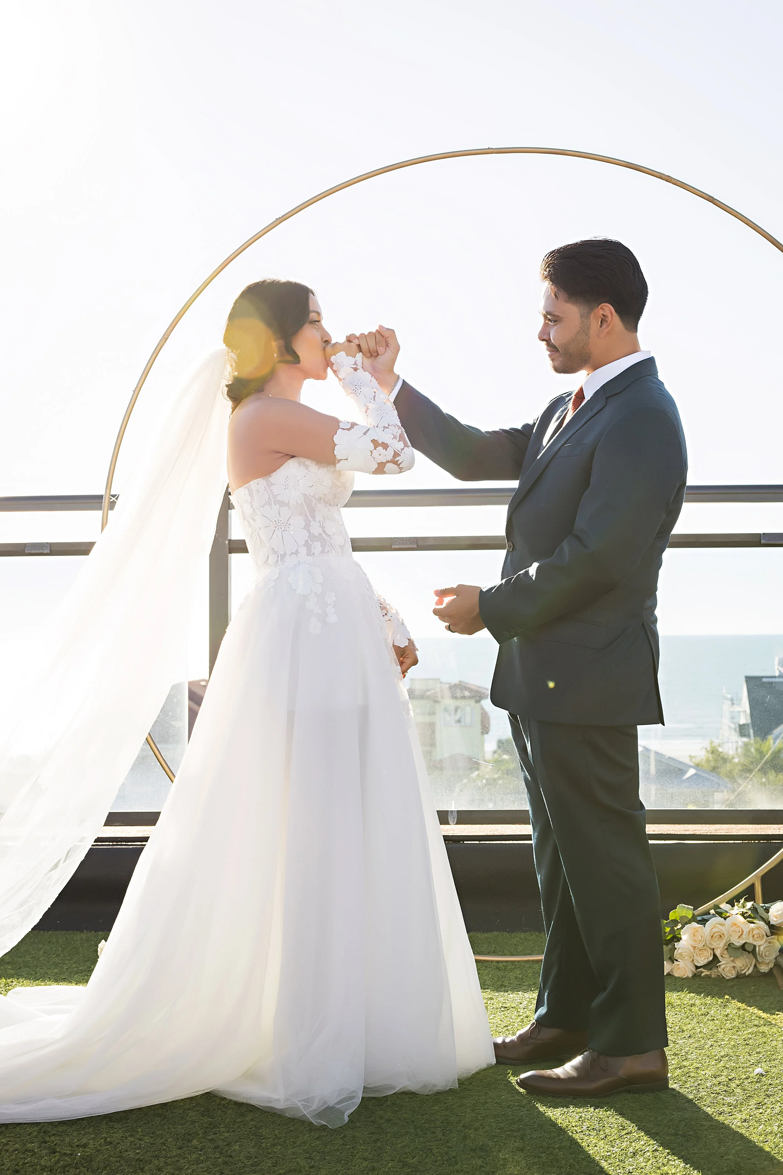 Groom in a navy suit spinning his bride in a white lace off-the-shoulder gown under a circular gold arch on a rooftop ceremony venue, with the city skyline and bright sky behind them.