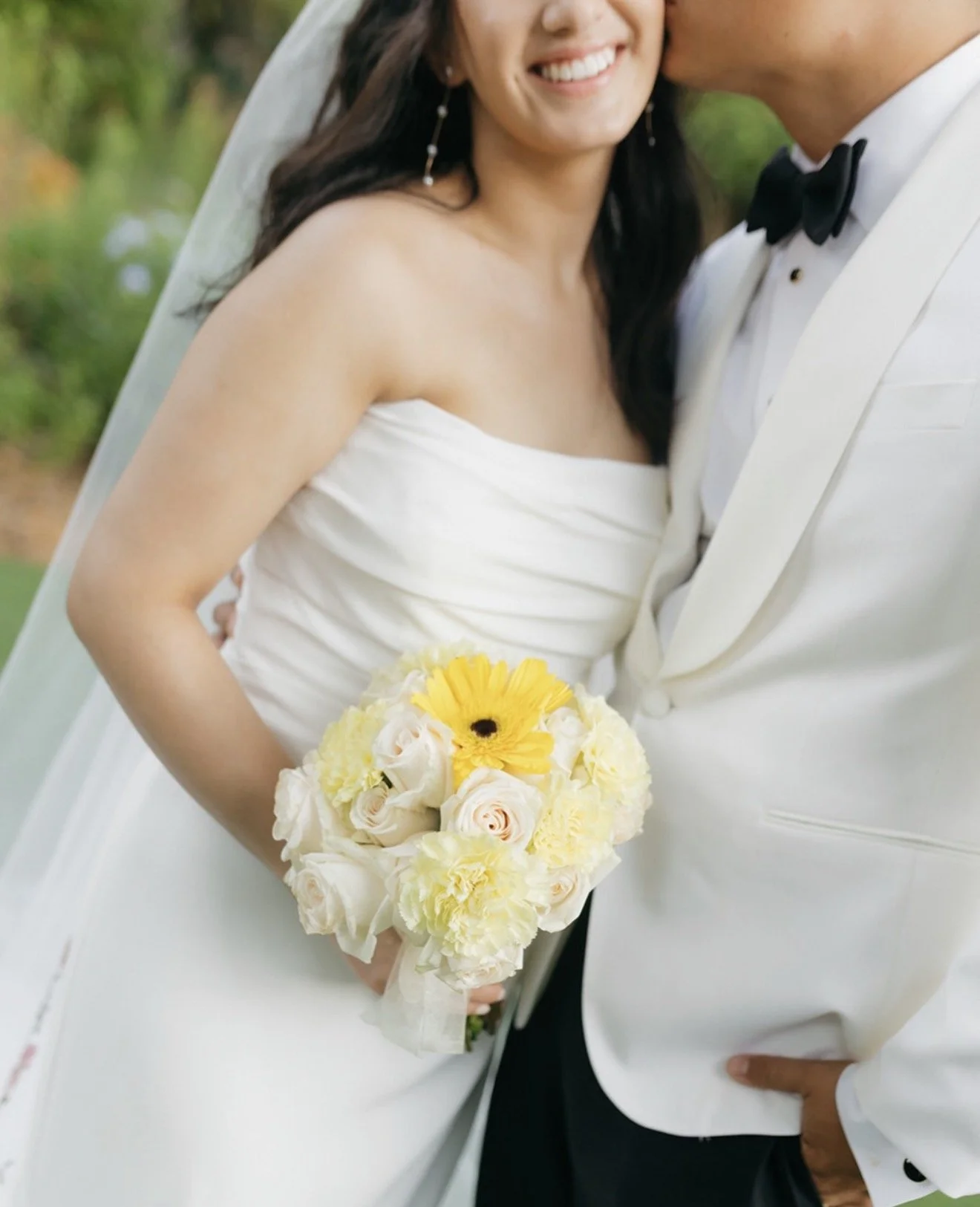 Groom in white tuxedo kissing bride's temple while she smiles and holds a yellow sunflower, white rose, and carnation bouquet at Sunken Gardens Tampa wedding by Peony Wedding Co.