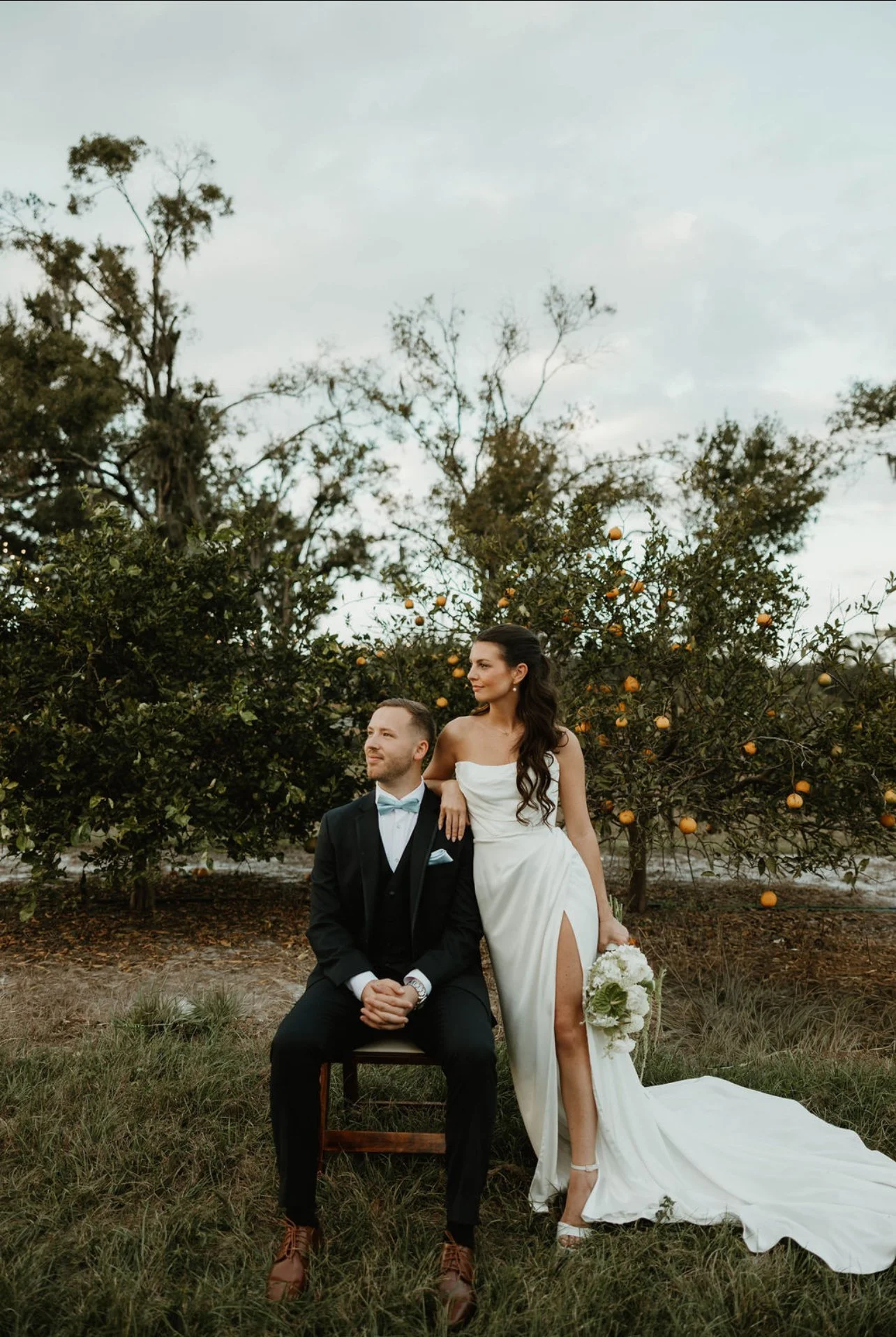 A newlywed couple stands on a grassy area near a pond surrounded by trees. The groom wears a black suit and tie, while the bride wears a white wedding dress with a long train and holds a bouquet of flowers. They embrace and smile at the camera.