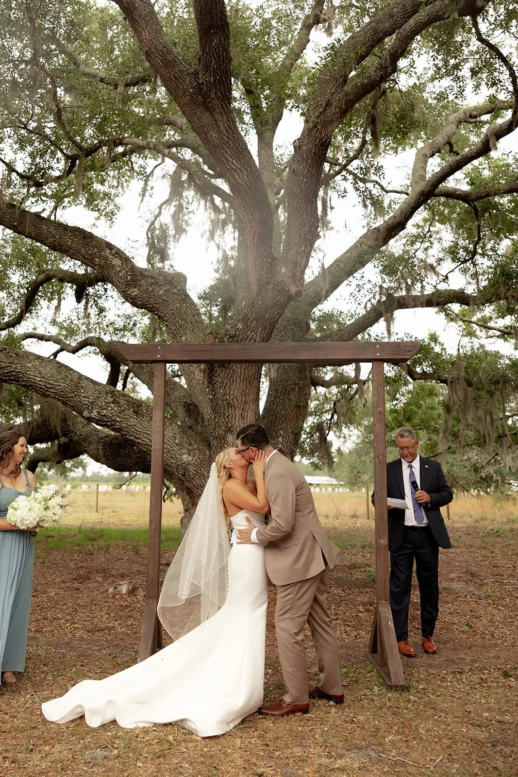 Bride and groom sharing first kiss at ceremony under the large live oak tree and wooden arch at Browns Grove Sarasota
