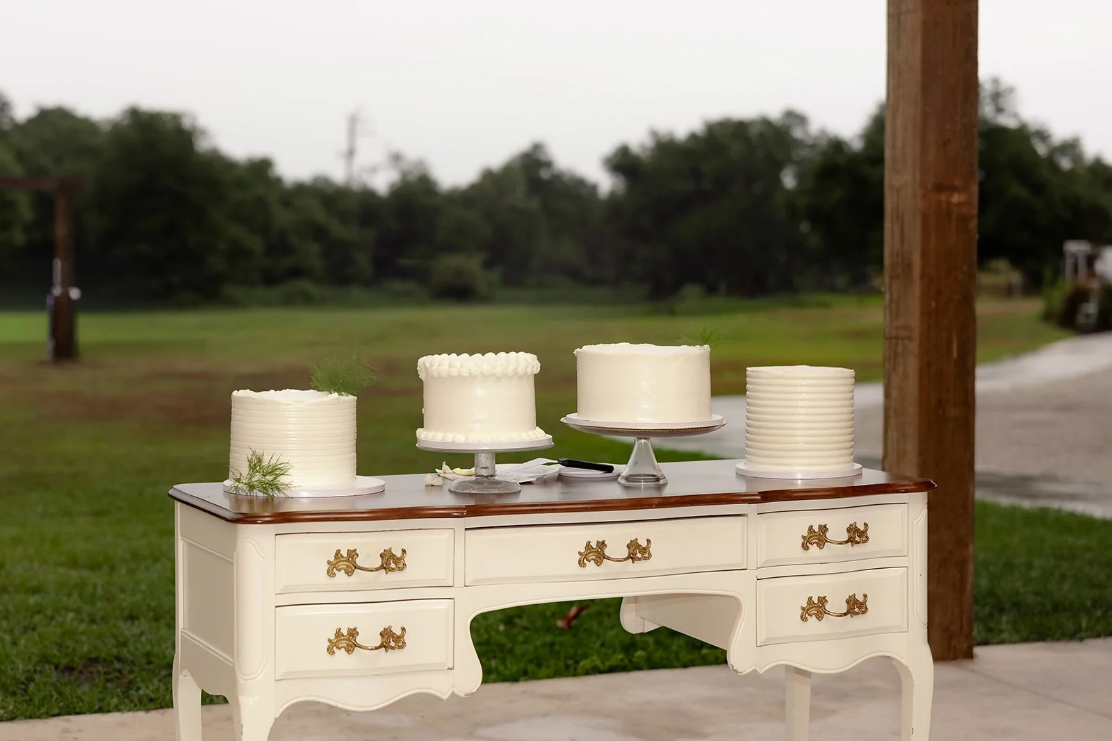 Four white wedding cakes in different styles displayed on a vintage white French provincial dresser outdoors at Browns Grove Sarasota
