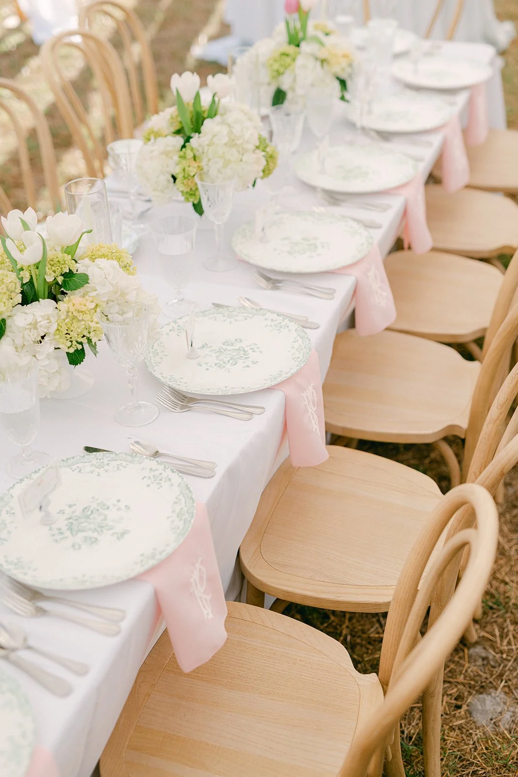 Wedding head table adorned with white hydrangea and tulip centerpieces, vintage green floral plates, crystal glassware, and blush pink monogrammed napkins draped over natural bentwood chairs at a Tampa wedding