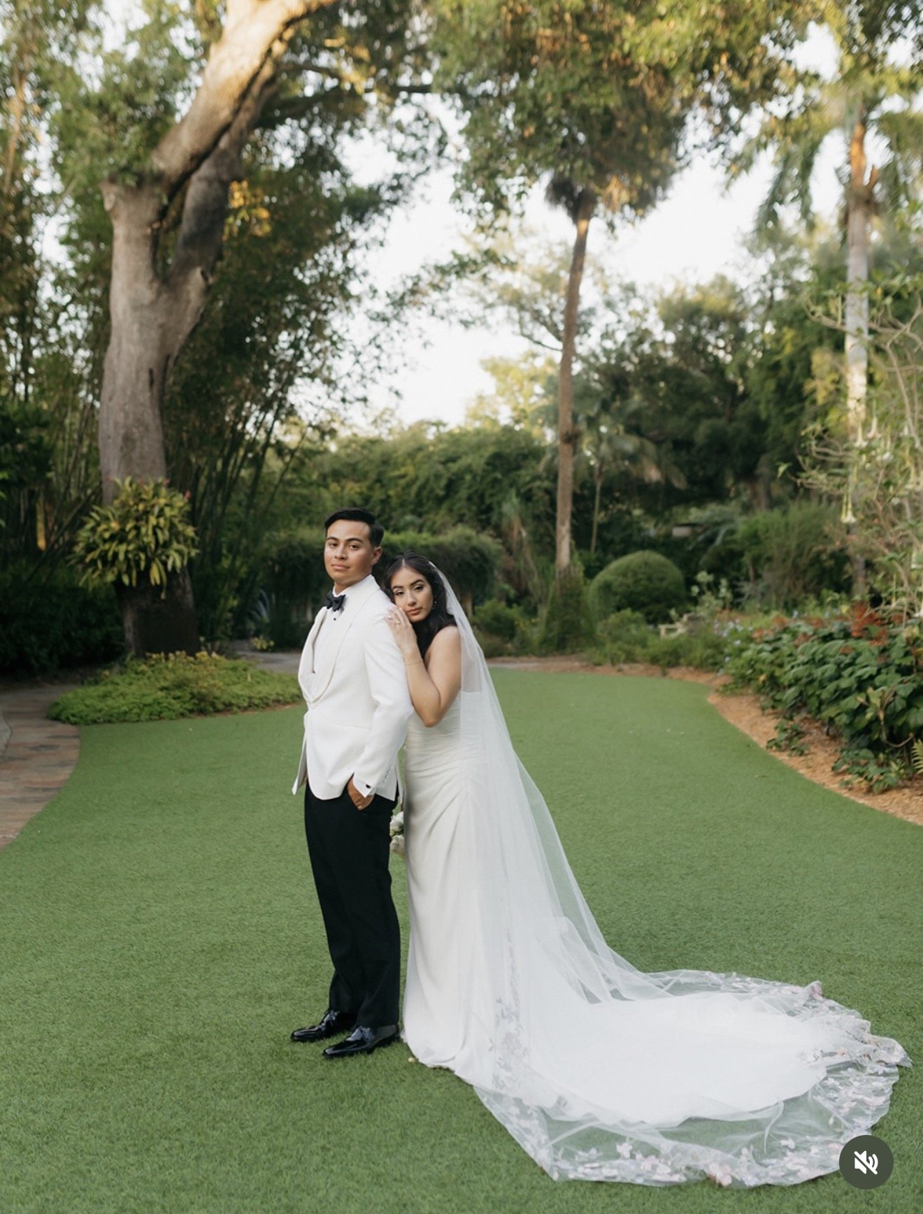 Bride in strapless white gown with cathedral-length lace-edged veil leaning against groom in white tuxedo on manicured lawn surrounded by tropical gardens and a large oak tree at Sunken Gardens Tampa, planned by Peony Wedding Co.