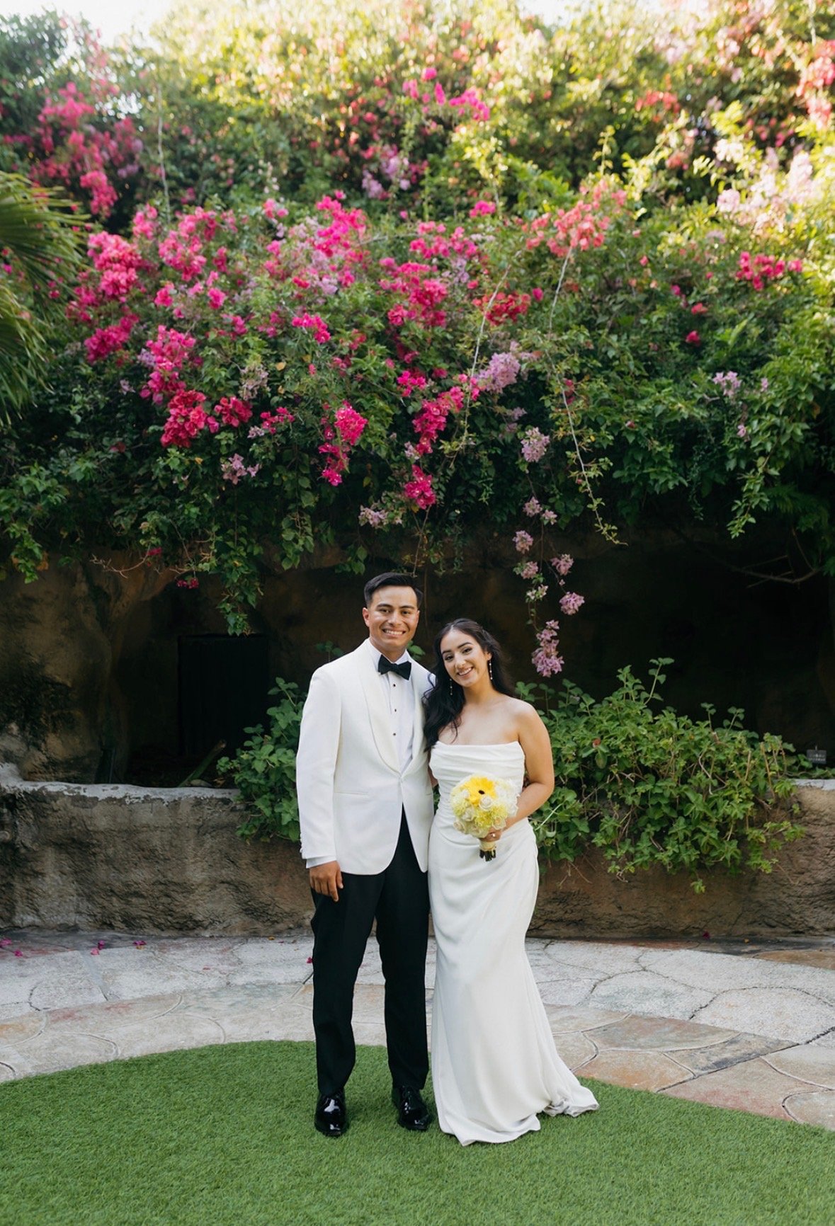 Bride and groom smiling together in front of a stone wall covered in cascading hot-pink bougainvillea at Sunken Gardens Tampa, Florida, planned by Peony Wedding Co.