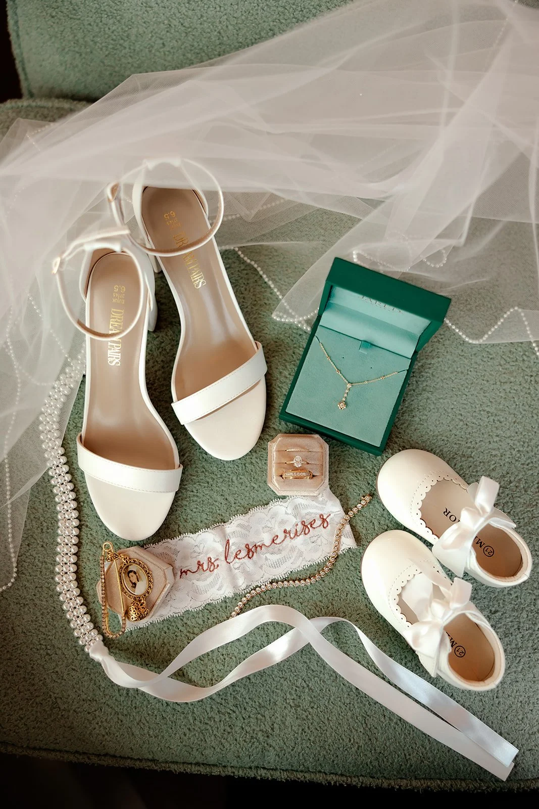 Bridal detail flat lay featuring white heeled sandals, Tiffany necklace, engagement ring, Mrs. Lesmerises garter, baby shoes, and pearl veil — Browns Grove Sarasota wedding