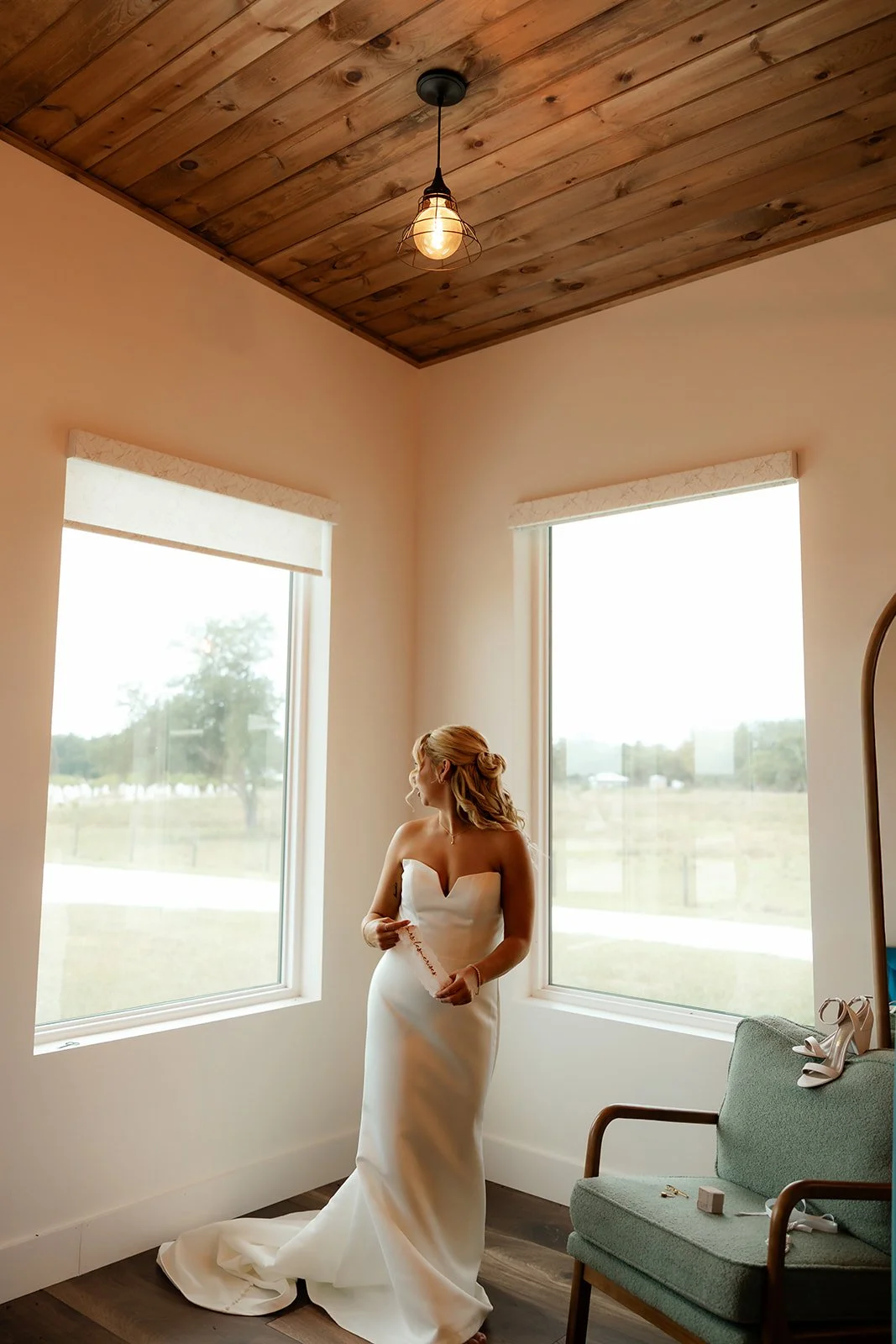 Bride in sweetheart wedding gown looking out the window of the Browns Grove Sarasota bridal suite, wood plank ceiling and pendant light