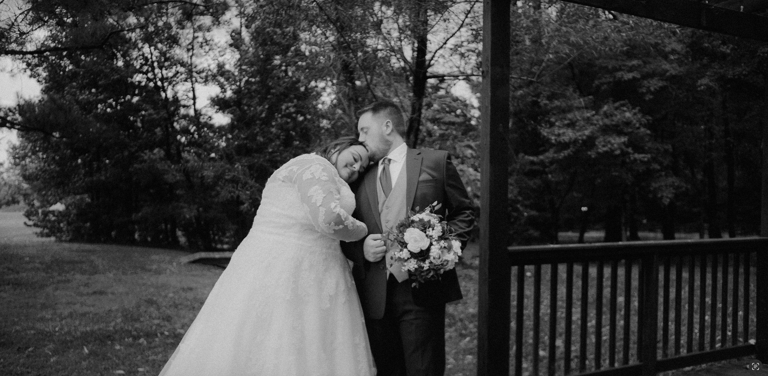 Black and white portrait of a plus-size bride in a long-sleeved lace wedding gown leaning her head on her groom's shoulder while he holds her bouquet of mixed florals, standing on a wooden deck surrounded by trees.