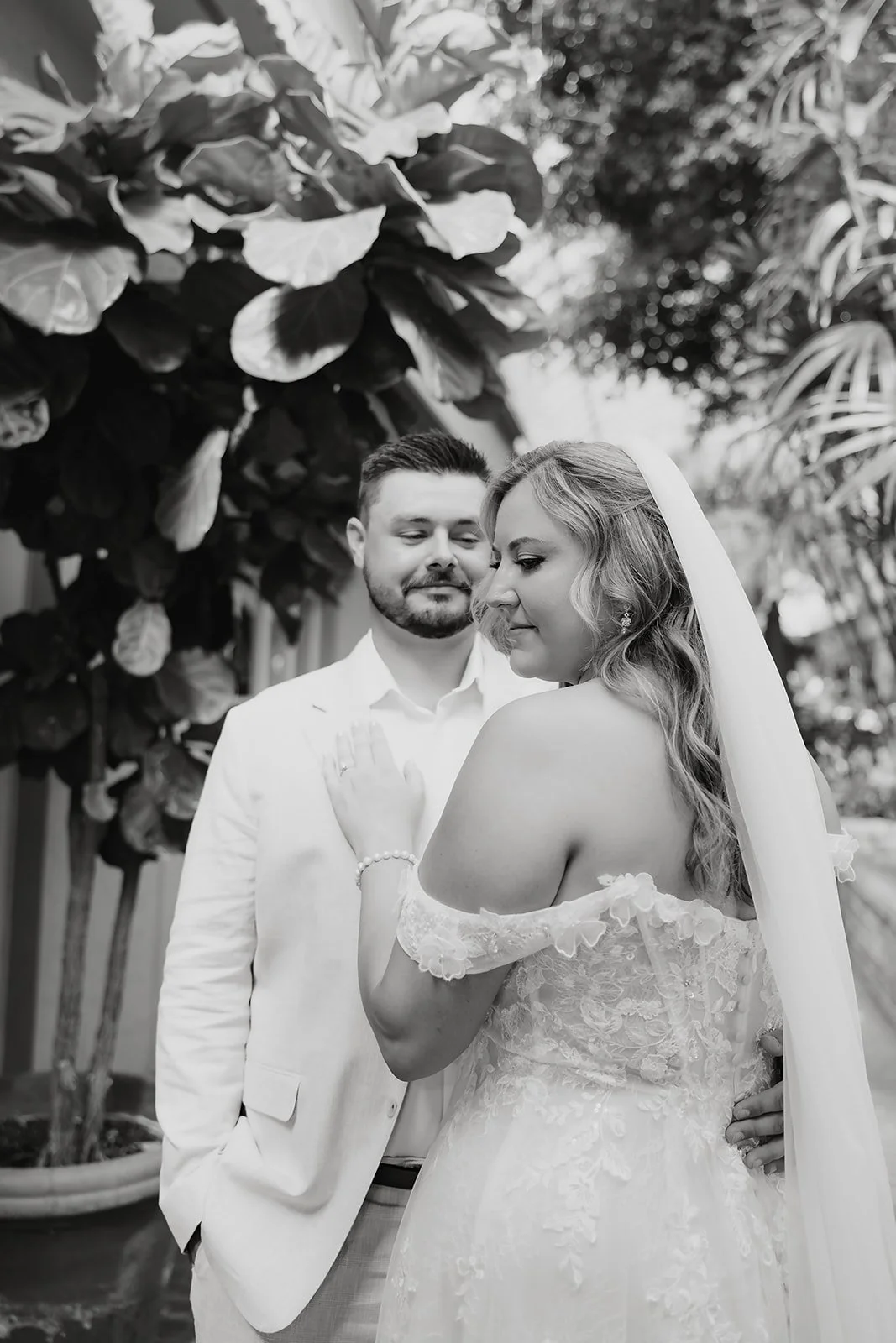 Black and white close-up of a bride in a lace wedding dress being gently kissed or embraced, with a soft bokeh background of trees and outdoor light.