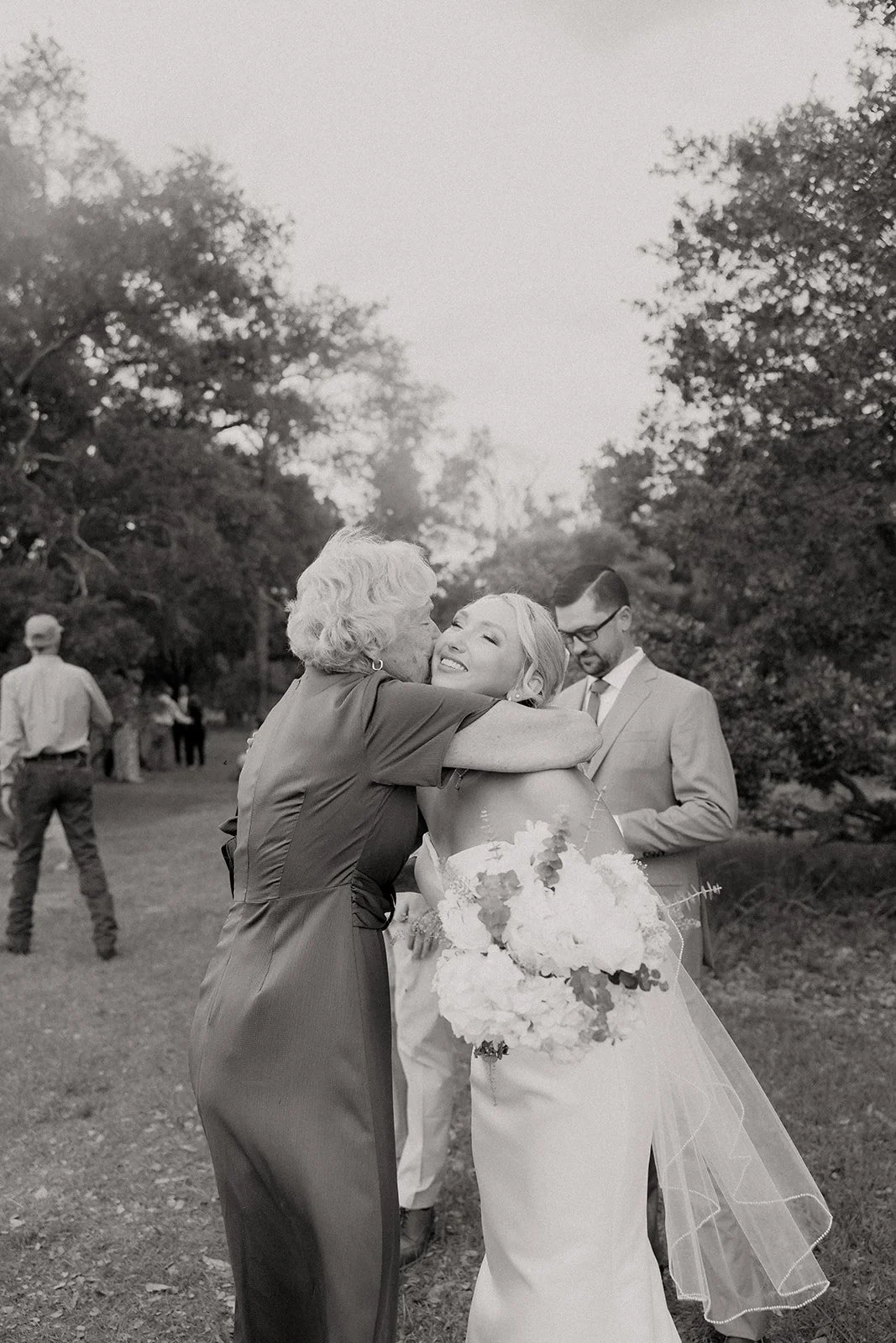 Black and white photo of bride being embraced by grandmother after ceremony at Browns Grove Sarasota, groom visible in background