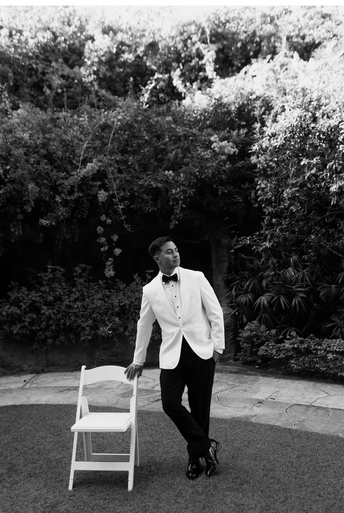 Black and white portrait of groom in white dinner jacket and black bow tie leaning against a white folding chair in the tropical garden at Sunken Gardens Tampa, planned by Peony Wedding Co.