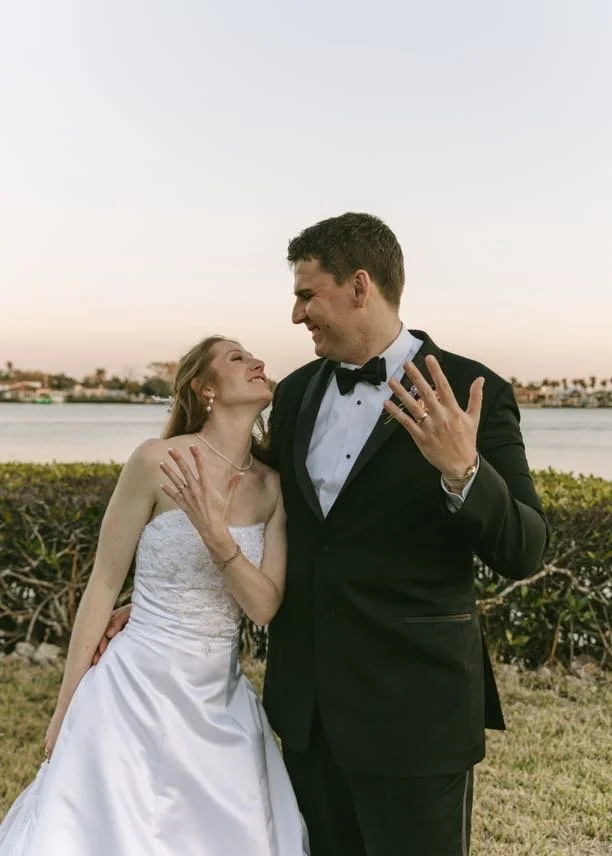Bride and groom laughing and looking at each other while showing their wedding rings, set against a glowing Florida waterfront at golden hour — a candid and joyful couples portrait from a Tampa Bay wedding