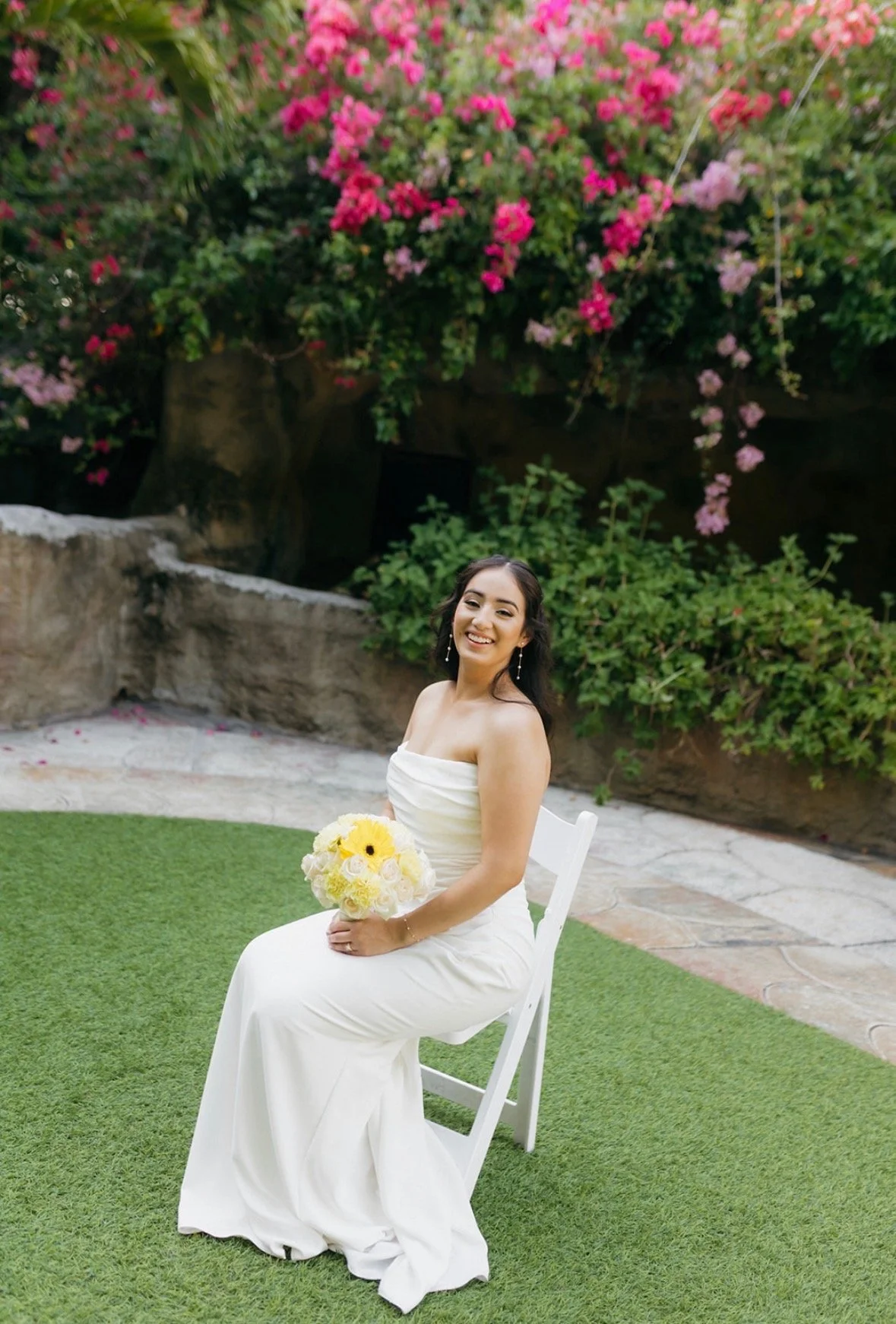 Smiling bride in strapless white gown seated on a white folding chair holding a yellow and white bouquet, with a lush wall of pink and magenta bougainvillea behind her at Sunken Gardens Tampa, by Peony Wedding Co.