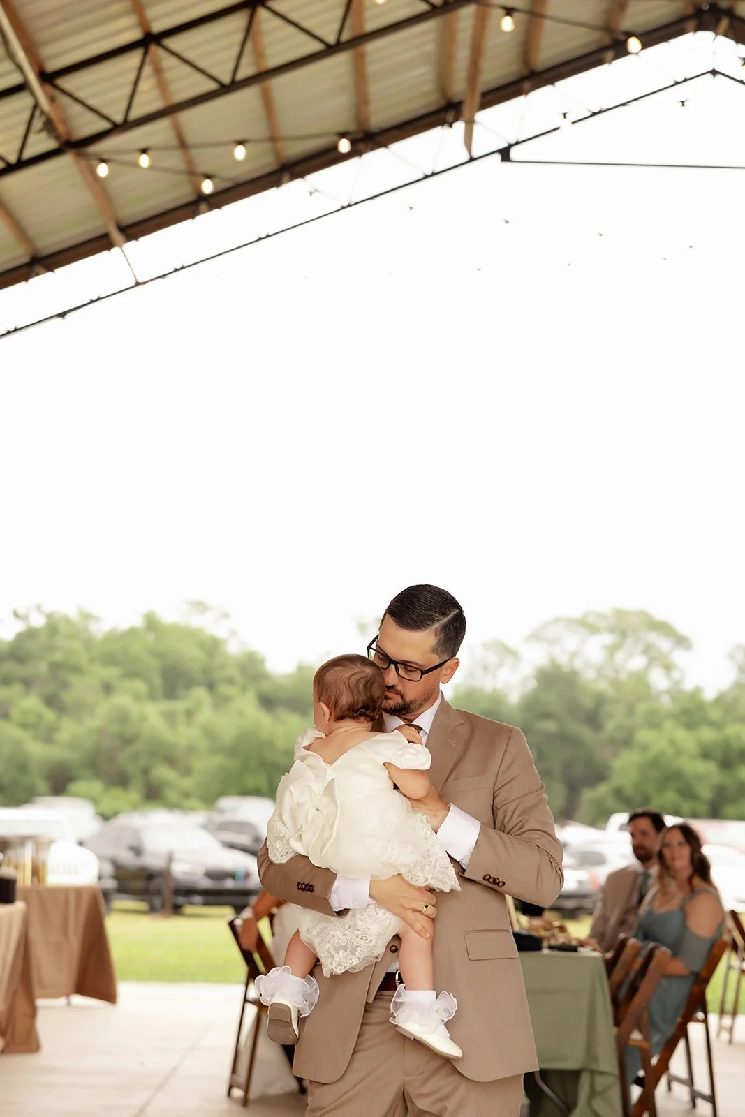 Groom holding baby daughter in lace dress during reception at Browns Grove Sarasota pavilion with string lights