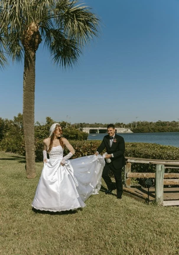 Bride holding up her wedding gown while the groom helps carry her train, walking together past a tall palm tree by a Florida waterfront with a wooden fence and calm water behind them — a tropical and candid couples portrait from a Tampa Bay wedding