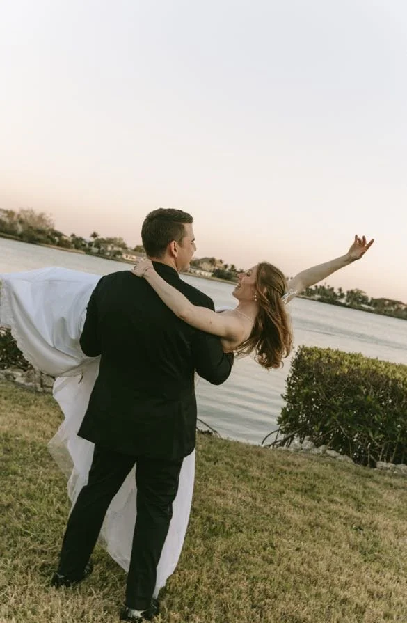Groom carrying his bride in his arms as she reaches her arm out and laughs, with the warm glow of sunset over Tampa Bay waterfront and tropical foliage behind them — a candid and romantic waterfront wedding portrait
