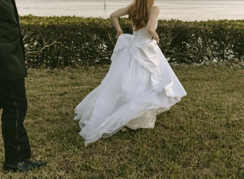Bride's flowing white layered ballgown from behind as she walks across a lawn by a Florida waterfront, with a groom partially visible beside her — a dreamy waterfront wedding moment from a Tampa Bay wedding