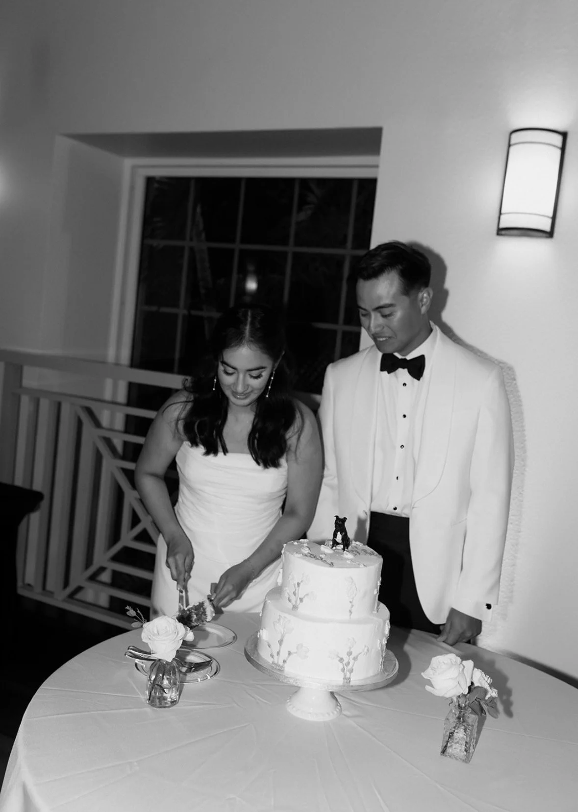 Black and white photo of bride and groom cutting a two-tier white wedding cake with floral buttercream detail and a dog cake topper at their Tampa wedding reception, planned by Peony Wedding Co.