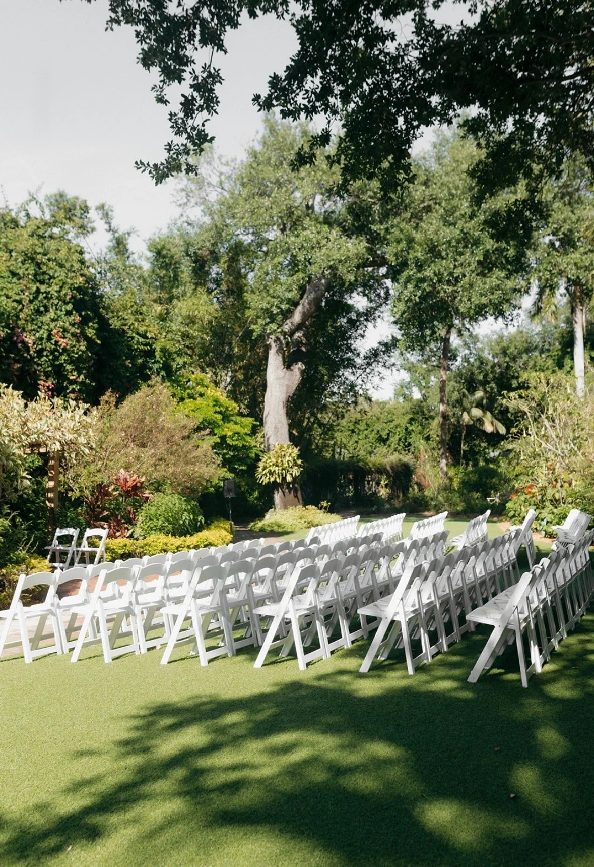 Rows of white folding chairs set up on a green lawn for an outdoor wedding ceremony at Sunken Gardens in Tampa, Florida, surrounded by mature oak trees and tropical gardens, planned by Peony Wedding Co.