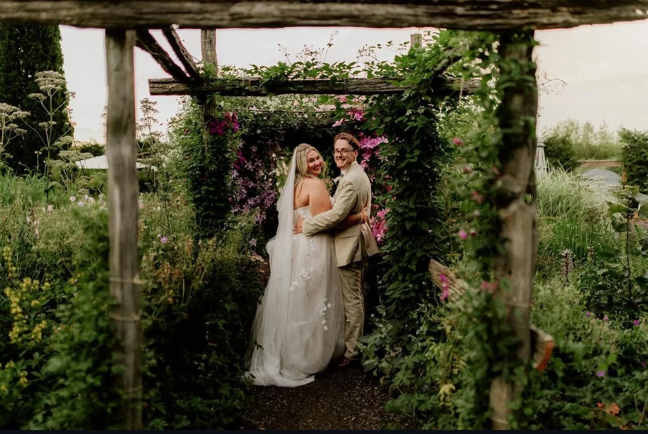 Bride in a flowing white wedding gown and groom in a tan suit embracing and laughing together under a rustic wooden pergola covered in lush greenery and wildflowers.