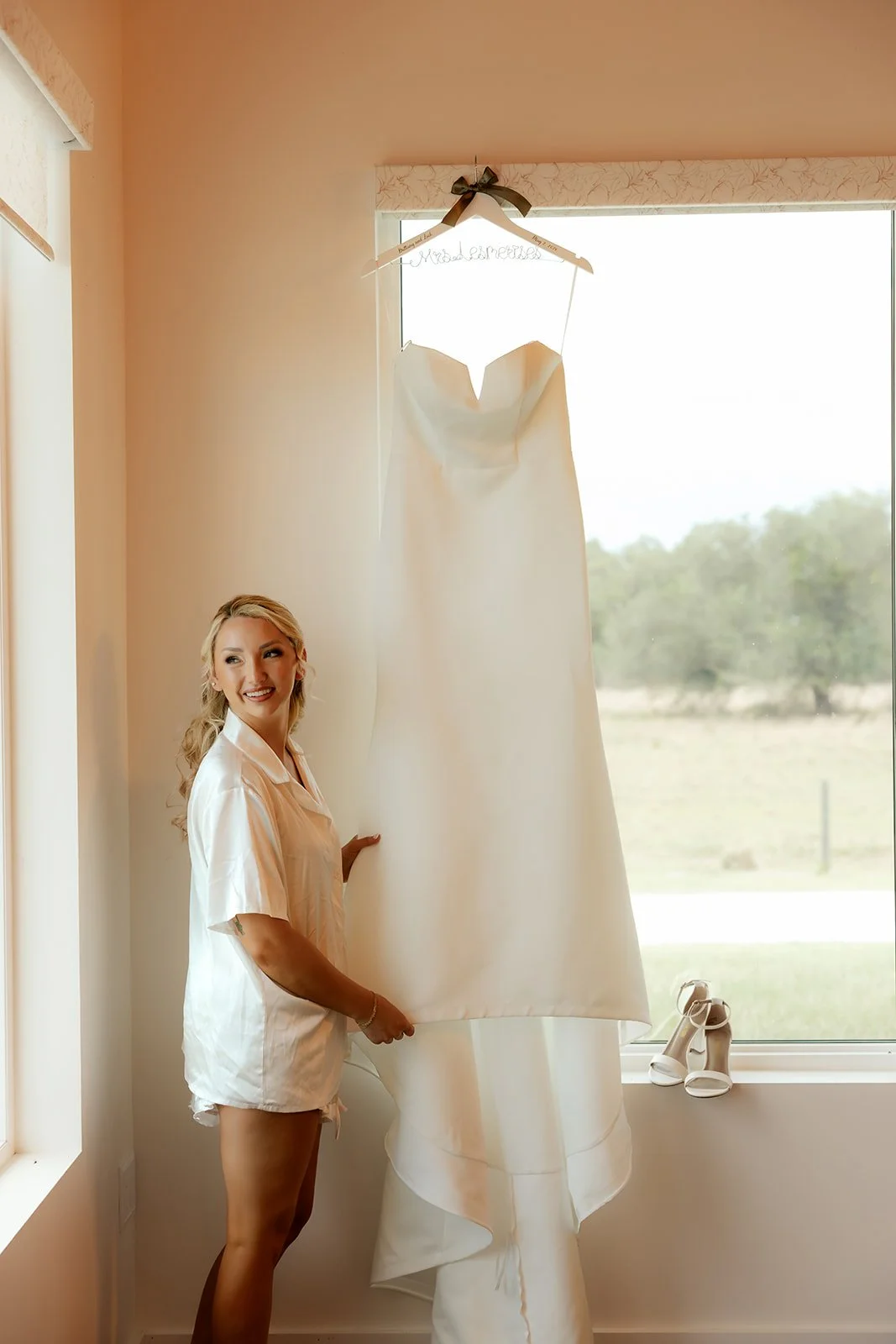 Smiling bride in white robe standing beside her wedding dress hanging in the window at Browns Grove Sarasota bridal suite